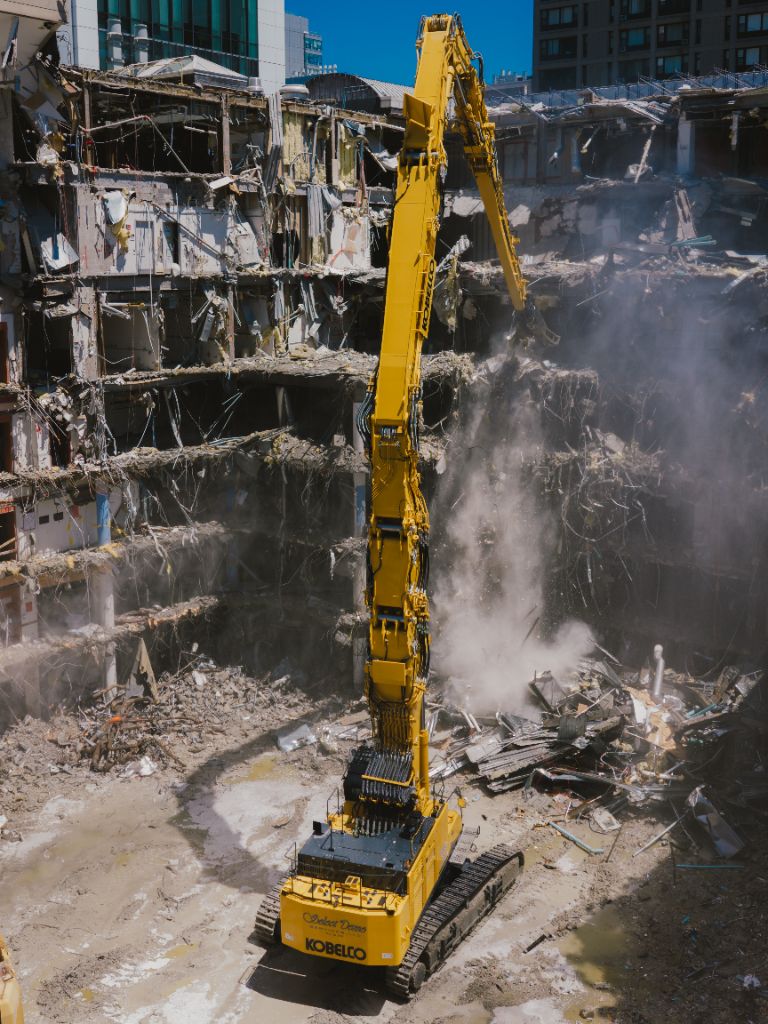 Kobelco high-reach excavator demolishing multi-storey building on active job site